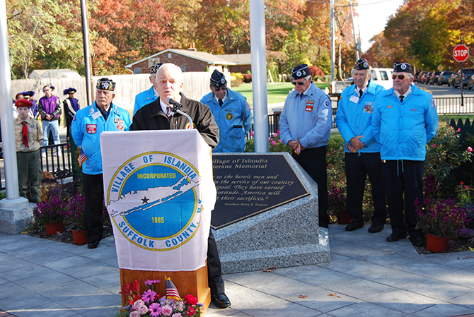 Mayor Allan M. Dorman (behind podium) speaks to the attendees at the Veterans Day ceremony at the Veterans Memorial Triangle on November 8 as members of the Central Long Island chapter of the Korean War Veterans Association look on.